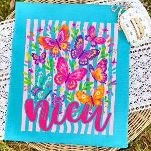 May include: A bright turquoise t-shirt featuring a graphic print of colorful butterflies in shades of pink, orange, purple, and teal. The design includes vertical light pink stripes, small star accents, and the word "Nicu" in large pink script.