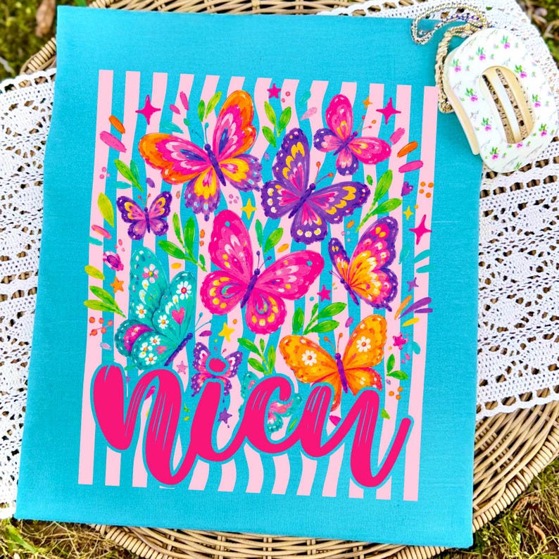 May include: A bright turquoise t-shirt featuring a graphic print of colorful butterflies in shades of pink, orange, purple, and teal. The design includes vertical light pink stripes, small star accents, and the word "Nicu" in large pink script.