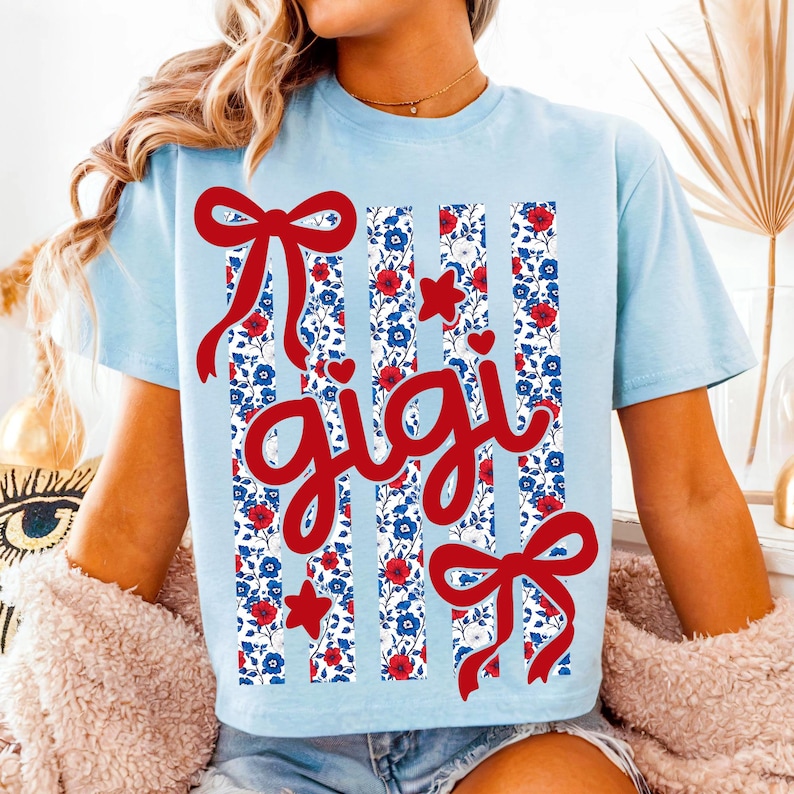 Op de afbeelding: Lichtblauw T-shirt met een rode strik en het woord "gigi" in rood schrift. Het shirt heeft verticale strepen met een bloemenpatroon in rood, wit en blauw, met rode harten en sterren.