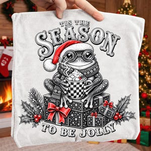 Puede incluir: Una camiseta blanca con una ilustraci&oacute;n en blanco y negro de una rana con gorro de Pap&aacute; Noel y gafas, sosteniendo una taza a cuadros con malvaviscos. El texto dice 'TIS THE SEASON TO BE JOLLY'. Bayas de acebo rojas y lazos decoran el dise&ntilde;o.