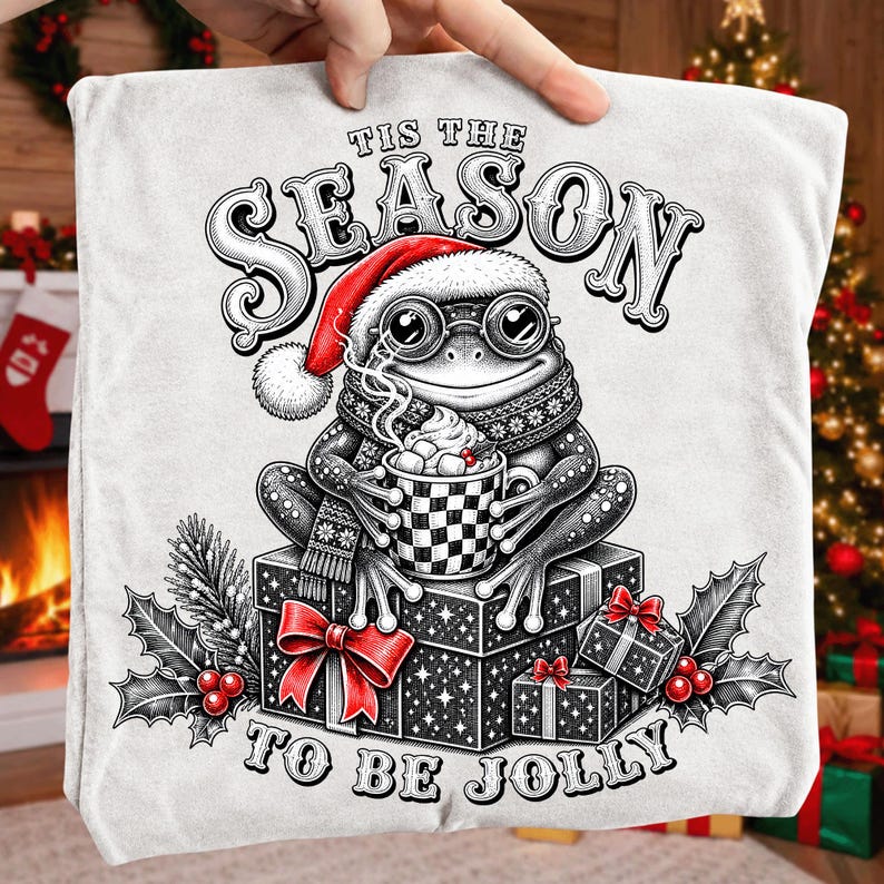 Puede incluir: Una camiseta blanca con una ilustraci&oacute;n en blanco y negro de una rana con gorro de Pap&aacute; Noel y gafas, sosteniendo una taza a cuadros con malvaviscos. El texto dice 'TIS THE SEASON TO BE JOLLY'. Bayas de acebo rojas y lazos decoran el dise&ntilde;o.
