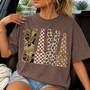 May include: A brown short-sleeved t-shirt featuring the word MAMA in large block letters. Each letter has a different pattern: camouflage, brown and white checkerboard, leopard print, and brown with white polka dots, accented by two bows.