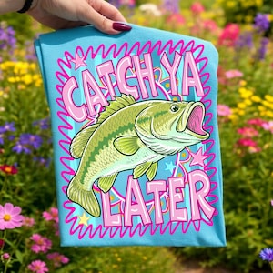 Op de afbeelding: Lichtblauw t-shirt met een grote groene en gele vis. De tekst "CATCH YA LATER" is roze, omlijnd door een golvend blauw ontwerp. Sterren en een kroon zijn ook in het ontwerp opgenomen.