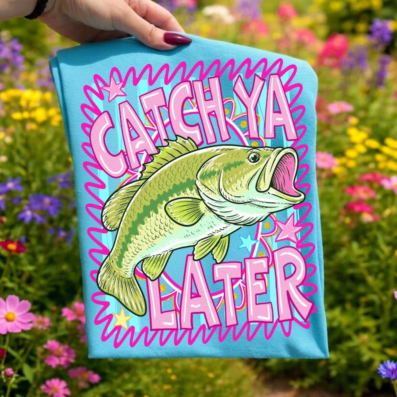 Op de afbeelding: Lichtblauw t-shirt met een grote groene en gele vis. De tekst "CATCH YA LATER" is roze, omlijnd door een golvend blauw ontwerp. Sterren en een kroon zijn ook in het ontwerp opgenomen.