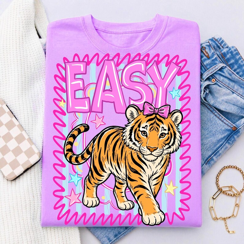 May include: A lilac t-shirt with a cartoon tiger wearing a pink bow. The word "EASY" is in pink bubble letters above the tiger, surrounded by a pink and blue striped border with stars.