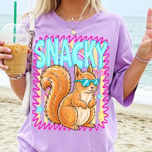 May include: A light purple t-shirt featuring a cartoon squirrel wearing blue sunglasses. The word "SNACKY" is in large, turquoise letters above the squirrel. The design includes stars and a pink zig-zag border.