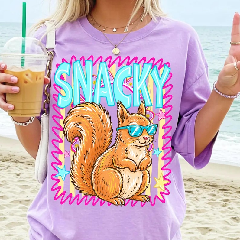 May include: A light purple t-shirt featuring a cartoon squirrel wearing blue sunglasses. The word "SNACKY" is in large, turquoise letters above the squirrel. The design includes stars and a pink zig-zag border.