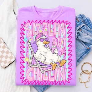 Op de afbeelding: Een lichtpaars t-shirt met een cartoon eend met een hartvormige zonnebril en een roze pet, ontspannen in een strandstoel. Het shirt heeft de tekst "Sizzlin' But Chillin'" in roze en wit, met een golvende rand.