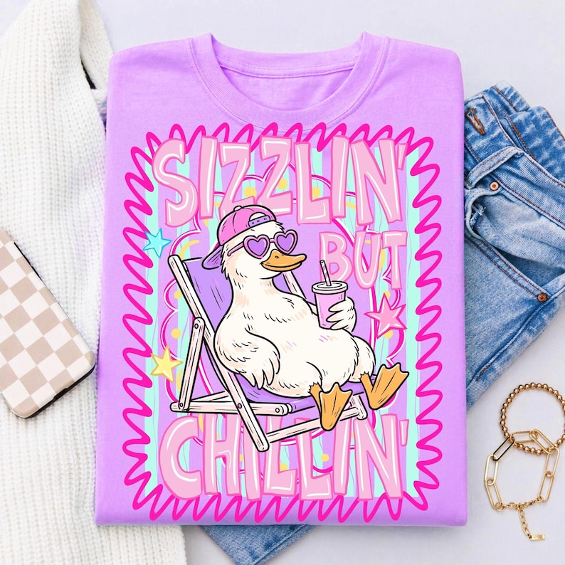 Op de afbeelding: Een lichtpaars t-shirt met een cartoon eend met een hartvormige zonnebril en een roze pet, ontspannen in een strandstoel. Het shirt heeft de tekst "Sizzlin' But Chillin'" in roze en wit, met een golvende rand.