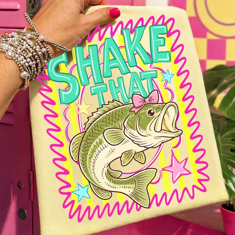 Op de afbeelding: Een lichtgele handdoek met de woorden "SHAKE THAT" in turquoise. In het midden staat een cartoon bas met een roze strik. Het ontwerp is omlijst door een roze golvende rand met sterren.