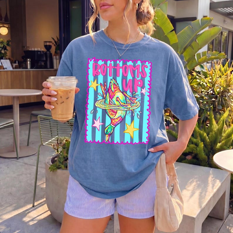 May include: Blue t-shirt featuring the text "Bottoms Up" and a cartoon duck diving into water. The shirt is worn with striped shorts. The person is holding a clear plastic cup with iced coffee.