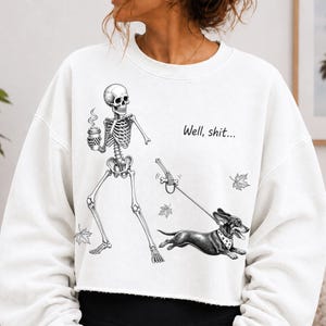 May include: A white long-sleeve sweatshirt featuring a black and white graphic of a skeleton holding a coffee cup and walking a dachshund on a leash. The text "Well, shit..." is printed above the dog, accompanied by small falling maple leaf illustrations.