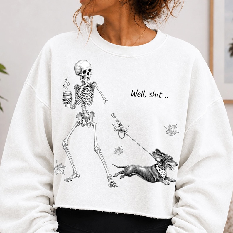 May include: A white long-sleeve sweatshirt featuring a black and white graphic of a skeleton holding a coffee cup and walking a dachshund on a leash. The text "Well, shit..." is printed above the dog, accompanied by small falling maple leaf illustrations.