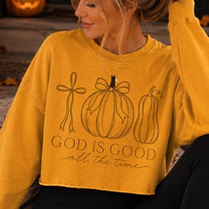 K&ouml;nnte beinhalten: Ein senfgelbes, bauchfreies Langarm-Sweatshirt mit einer schwarzen Strichzeichnung, die ein Kreuz, einen K&uuml;rbis mit Schleife und einen Flaschenk&uuml;rbis zeigt. Unter den Illustrationen steht der Text "GOD IS GOOD all the time" in Serif- und Schreibschrift.