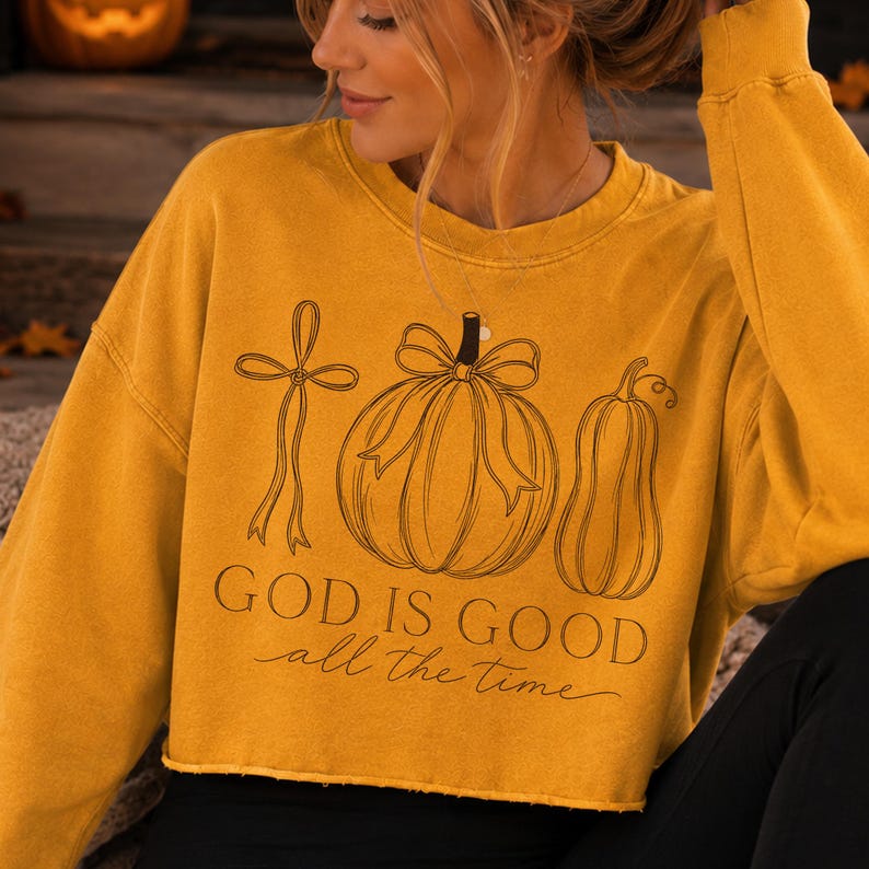 K&ouml;nnte beinhalten: Ein senfgelbes, bauchfreies Langarm-Sweatshirt mit einer schwarzen Strichzeichnung, die ein Kreuz, einen K&uuml;rbis mit Schleife und einen Flaschenk&uuml;rbis zeigt. Unter den Illustrationen steht der Text "GOD IS GOOD all the time" in Serif- und Schreibschrift.
