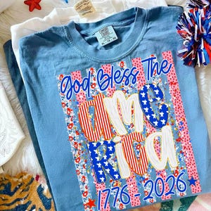 Puede incluir: Una camiseta azul claro con la frase "God Bless The America 1776-2026" en un dise&ntilde;o patri&oacute;tico. El dise&ntilde;o incorpora rayas rojas y blancas, estrellas azules y estampados florales. La camiseta es talla XL (ES 46-48).
