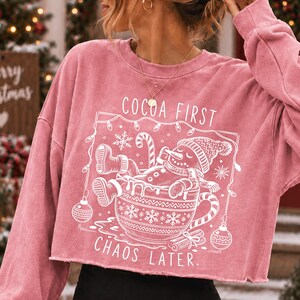 Op de afbeelding: Een oudroze cropped sweatshirt met lange mouwen en een witte grafische print van een sneeuwpop die ontspant in een grote mok. De tekst luidt COCOA FIRST CHAOS LATER, omringd door sneeuwvlokken, kerstverlichting en zuurstokken in een kader.