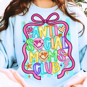 K&ouml;nnte beinhalten: Hellblaues Sweatshirt mit einem farbenfrohen Aufdruck &bdquo;ANTI SOCIAL MOMS CLUB&ldquo;. Das Design umfasst Smileys, Herzen, Blumen und einen Blitz, alles schwarz umrandet und auf einem rosa Band platziert.