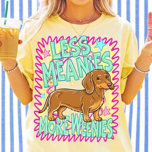 May include: Yellow t-shirt with a brown dachshund illustration and the text "LESS MEANIES MORE WEENIES" in a turquoise and pink design. The shirt has a crew neck and short sleeves.