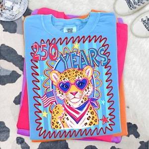 Op de afbeelding: Een stapel kleurrijke t-shirts. Het bovenste shirt is lichtblauw met een afbeelding van een luipaard met een hartvormige zonnebril en een Amerikaanse vlag. De tekst "250 YEARS" staat boven de luipaard. Andere shirts zijn roze, oranje en paars.