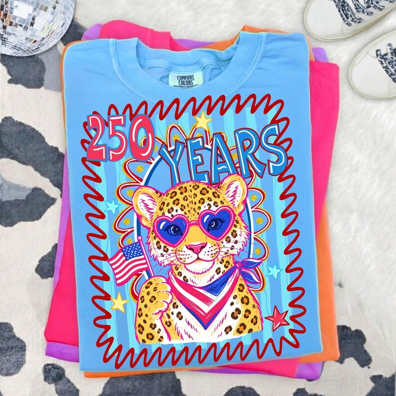 Op de afbeelding: Een stapel kleurrijke t-shirts. Het bovenste shirt is lichtblauw met een afbeelding van een luipaard met een hartvormige zonnebril en een Amerikaanse vlag. De tekst "250 YEARS" staat boven de luipaard. Andere shirts zijn roze, oranje en paars.