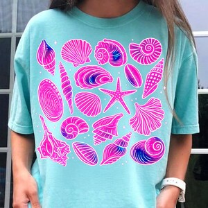 Pu&ograve; includere: Una maglietta azzurra con illustrazioni di conchiglie rosa e viola. Il design presenta varie forme di conchiglie, una stella marina e stelle, creando un'estetica estiva e a tema spiaggia.