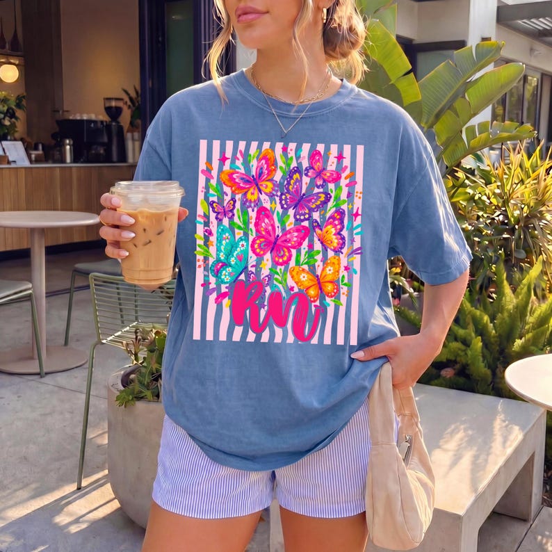 May include: A blue short-sleeve t-shirt featuring a graphic of colorful butterflies in shades of pink, orange, yellow, and teal, set against a background of vertical light pink and white stripes with the cursive pink text RN printed below.