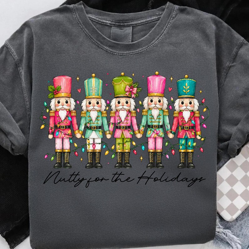 Op de afbeelding: Een antracietgrijs t-shirt met een afbeelding van vijf notenkrakers in roze, groene en rode uniformen die elkaars hand vasthouden, omringd door kleurrijke kerstverlichting. De tekst onder de figuren luidt Nutty for the Holidays.