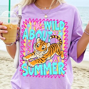 K&ouml;nnte beinhalten: Ein lila T-Shirt mit einem Tiger-Motiv und dem Schriftzug "WILD ABOUT SUMMER" in T&uuml;rkis. Der Tiger ist orange und schwarz, vor einem rosa und gelben Hintergrund mit Sternen. Das T-Shirt hat kurze &Auml;rmel und einen Rundhalsausschnitt.