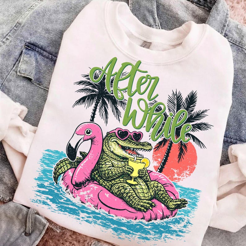 Pu&ograve; includere: Felpa bianca con un coccodrillo dei cartoni animati con occhiali da sole a forma di cuore, che tiene in mano un drink e galleggia su un galleggiante a forma di fenicottero rosa. Il testo "After While" &egrave; scritto sopra il coccodrillo. Palme e un tramonto sullo sfondo.