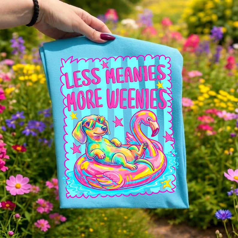 May include: A light blue t-shirt featuring a colorful graphic of a dachshund wearing heart-shaped sunglasses while lounging on a pink flamingo pool float. The text reads LESS MEANIES MORE WEENIES in bright pink letters, surrounded by stars.