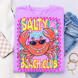 May include: A lilac t-shirt with a cartoon crab wearing sunglasses and a pearl necklace. The shirt features the words "SALTY MOMS BEACH CLUB" in yellow and turquoise lettering, with pink accents and stars.