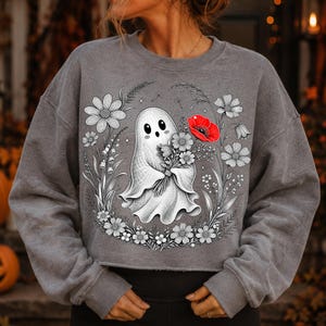 K&ouml;nnte beinhalten: Ein anthrazitfarbenes Sweatshirt mit Rundhalsausschnitt und einem Grafikdruck eines wei&szlig;en Geistes, der einen Blumenstrau&szlig; h&auml;lt. Das Design zeigt schwarz-wei&szlig;e Strichzeichnungen von Blumen um den Geist herum, mit einer einzelnen leuchtend roten Mohnblume.