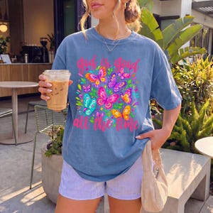 May include: A blue short-sleeve t-shirt featuring a colorful graphic of butterflies and green leaves with the pink text "god is good all the time" printed on the front. The shirt is paired with light blue and white pinstriped shorts.