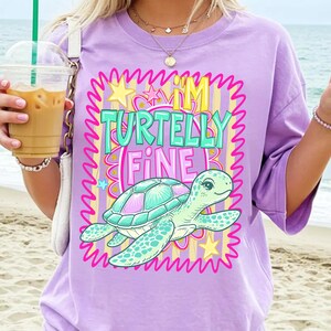 May include: A lilac-colored t-shirt with the text "I'M TURTELLY FINE" and a cartoon turtle graphic. The turtle has a teal and pink shell. The text is in teal and pink with a yellow crown above the "I'M".