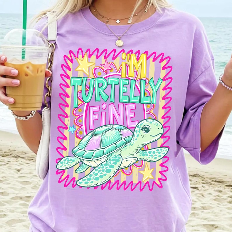 May include: A lilac-colored t-shirt with the text "I'M TURTELLY FINE" and a cartoon turtle graphic. The turtle has a teal and pink shell. The text is in teal and pink with a yellow crown above the "I'M".