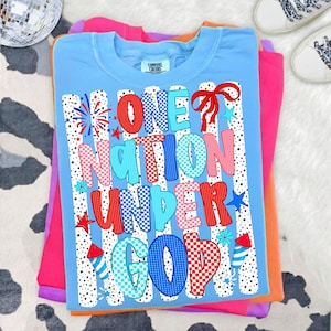 Puede incluir: Una camiseta Comfort Colors azul claro con el texto "ONE NATION UNDER GOD" en rojo, rosa y azul. El dise&ntilde;o incluye fuegos artificiales, estrellas y un lazo. La camiseta est&aacute; doblada encima de otras camisetas coloridas.