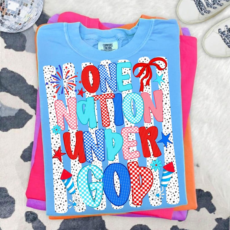 Puede incluir: Una camiseta Comfort Colors azul claro con el texto "ONE NATION UNDER GOD" en rojo, rosa y azul. El dise&ntilde;o incluye fuegos artificiales, estrellas y un lazo. La camiseta est&aacute; doblada encima de otras camisetas coloridas.