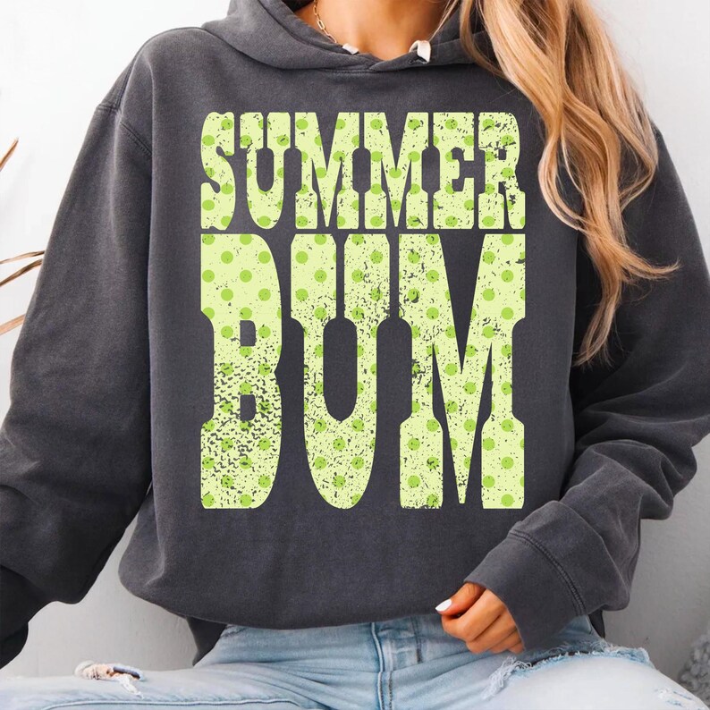 Puede incluir: Sudadera con capucha gris oscuro con la frase "SUMMER BUM" en letras grandes de color verde lima con lunares. La sudadera tiene una capucha con cord&oacute;n y mangas largas. El texto tiene un aspecto vintage.