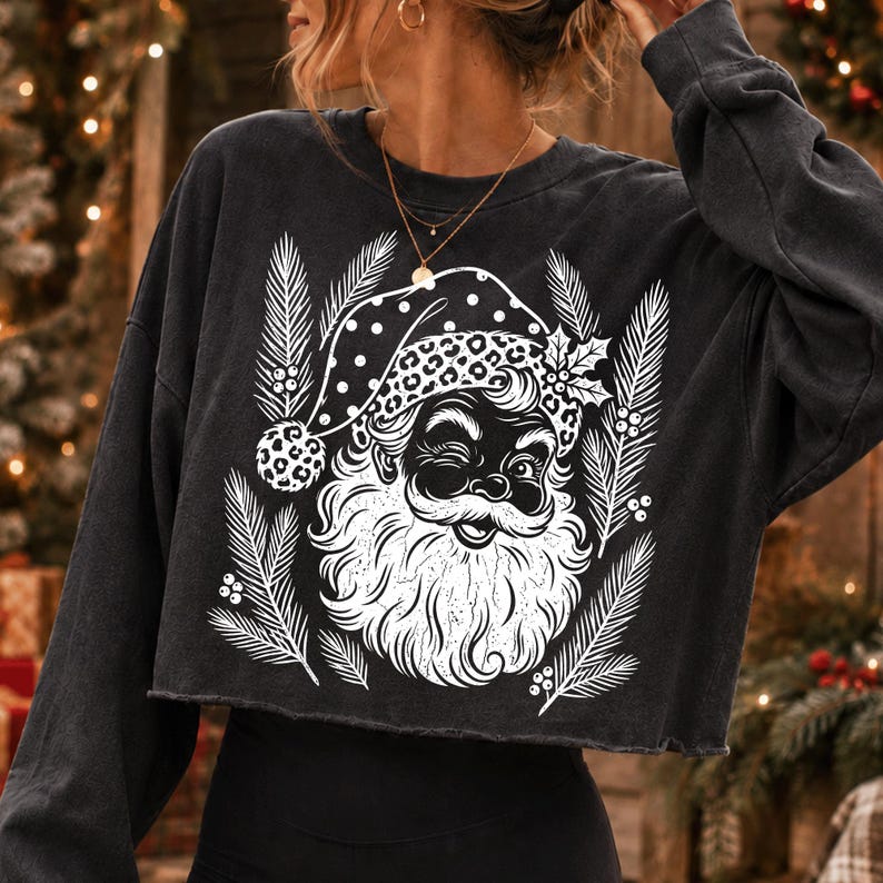 Puede incluir: Una sudadera corta de color gris carb&oacute;n y manga larga con un estampado gr&aacute;fico blanco de un Santa Claus gui&ntilde;ando un ojo, con un gorro de estampado de leopardo, rodeado de ramas de pino y bayas de acebo estilizadas en el centro.