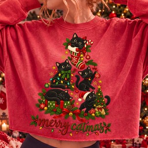 K&ouml;nnte beinhalten: Ein rotes Langarm-Sweatshirt mit einer Grafik von f&uuml;nf schwarzen Katzen, die in Form eines Weihnachtsbaums angeordnet sind. Die Katzen sind mit Ornamenten, Lichtern und einer Weihnachtsm&uuml;tze geschm&uuml;ckt. Der Text "merry catmas" steht unten in Gr&uuml;n.