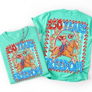 May include: Two turquoise t-shirts with a graphic design. The design features a cowgirl riding a horse with a lasso, surrounded by the text "250 YEARS OF FREEDOM" in red and blue. The shirts are made of a soft material.
