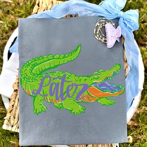 Pu&ograve; includere: Una borsa tote grigia con un design di alligatore verde con dettagli arancioni e viola. La parola "Patez" &egrave; scritta in corsivo viola sul corpo dell'alligatore. Un nastro azzurro e un fermaglio decorativo per capelli sono attaccati alla borsa.