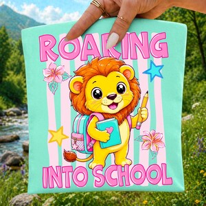 May include: A light mint green fabric square featuring a cartoon lion with a brown mane, wearing a pink backpack, holding a blue book and a yellow pencil. The text ROARING INTO SCHOOL is printed in pink. Pink vertical stripes decorate the background.
