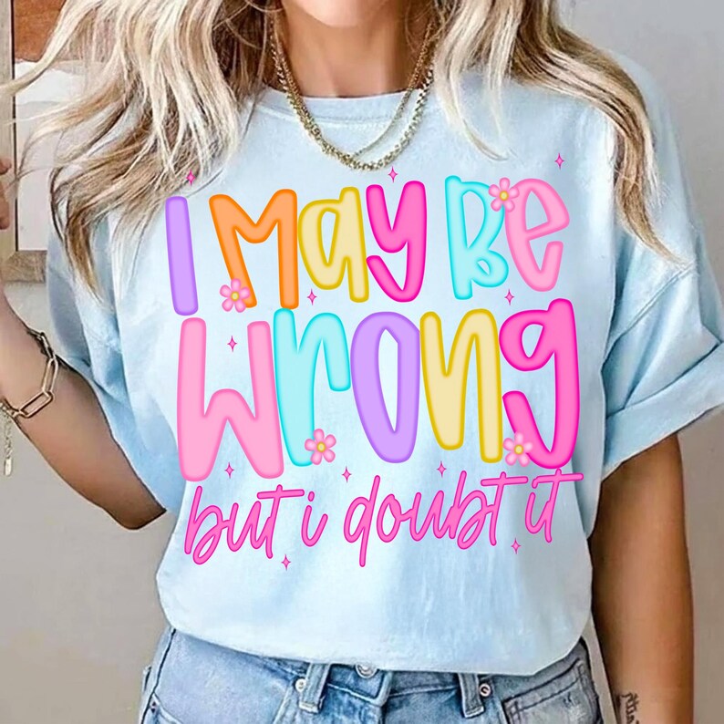 Op de afbeelding: Lichtblauw T-shirt met de tekst "I MAY BE WRONG but i doubt it" in kleurrijke letters. De tekst bevat oranje, roze, geel en blauw met kleine bloemaccenten. Het shirt heeft korte mouwen en een ronde hals.