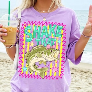 Op de afbeelding: Een lichtpaars T-shirt met de tekst "SHAKE THAT" in turquoise letters. Onder de tekst staat een cartoon baarsvis met een roze strik. Het ontwerp is omlijst door een roze zigzagrand en gele strepen.