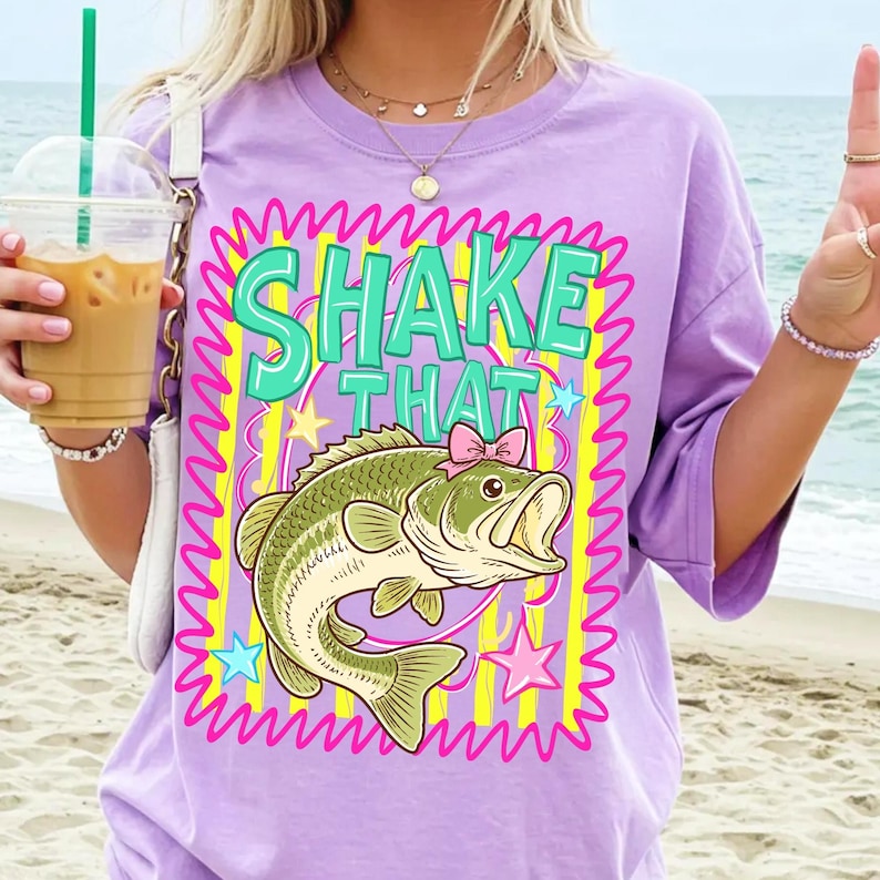 Op de afbeelding: Een lichtpaars T-shirt met de tekst "SHAKE THAT" in turquoise letters. Onder de tekst staat een cartoon baarsvis met een roze strik. Het ontwerp is omlijst door een roze zigzagrand en gele strepen.