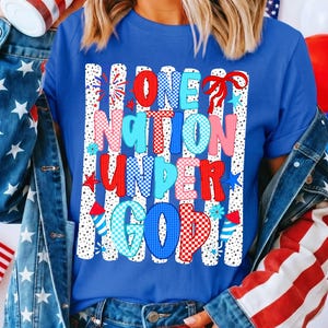 Puede incluir: Una camiseta azul con la frase "ONE NATION UNDER GOD" en letras rojas, azules y rosas. El dise&ntilde;o incluye fuegos artificiales, estrellas y detalles florales, sobre un fondo de rayas blancas con lunares negros. La camiseta tiene un tema patri&oacute;tico.