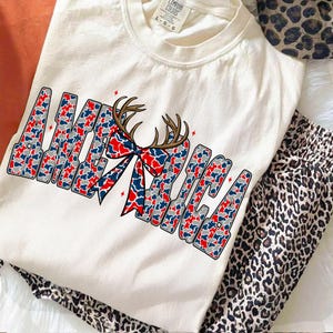 Pu&ograve; includere: T-shirt color crema con la scritta "AMERICA" in un motivo mimetico rosso, bianco e blu. Un fiocco e un motivo di corna sono centrati sopra la parola. La maglietta &egrave; abbinata a pantaloni con stampa leopardata.