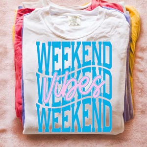 K&ouml;nnte beinhalten: Ein Stapel gefalteter T-Shirts in verschiedenen Farben, darunter Wei&szlig;, Rot, Lila und Gelb. Das oberste wei&szlig;e Shirt zeigt den Text "WEEKEND Vibes WEEKEND" in blauer und rosa Schrift.